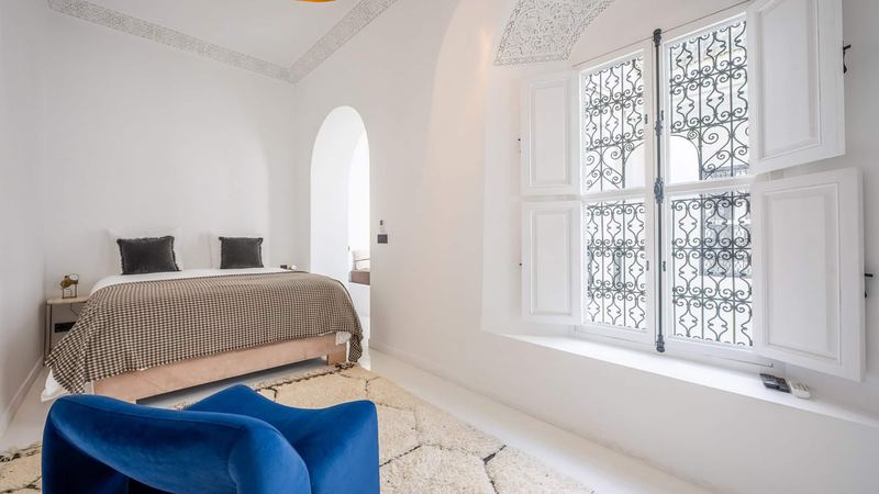 Bedroom 2: the queen bed under a houndstooth throw, a blue velvet armchair, a pale rug and a wrought-iron window with white shutters.