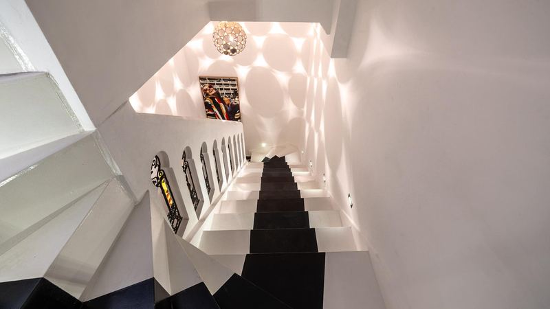 The stairs seen from above: white steps with a black runner, small wrought-iron windows and the light of a pendant lamp on the walls.