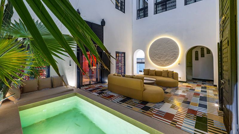 The courtyard at dusk, seen through palm leaves: the pool in front, the mustard sofas, the cement-tile floor and a lit white disc on the wall.