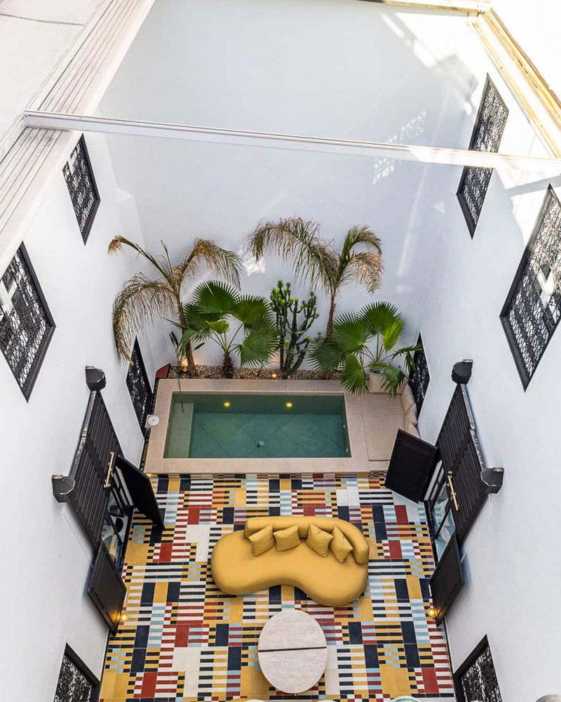 The courtyard seen from the top of the house: the pool, the palms, the mustard sofa and the cement tiles, between wrought-iron windows.