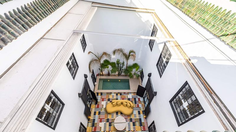The courtyard seen from the top of the house: the pool, the palms, the mustard sofa and the cement tiles, between wrought-iron windows.