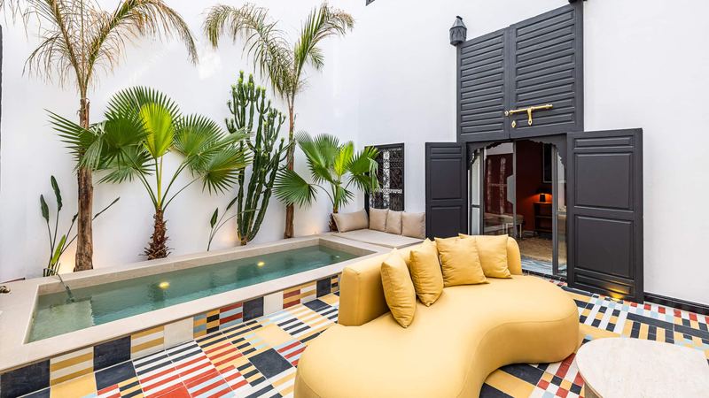 The courtyard in daylight: the private pool, a curved mustard sofa, palms and the multicoloured cement-tile floor; the black doors open onto the terracotta-pink sitting room.