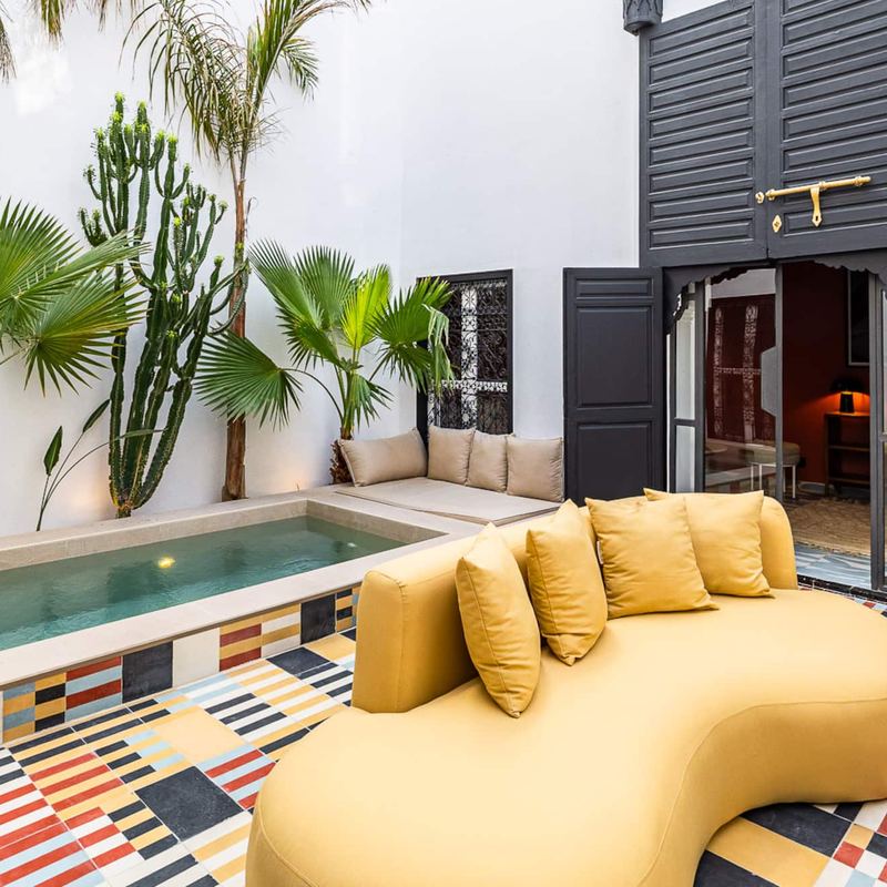Le patio en plein jour : la piscine privée, un canapé courbe couleur moutarde, des palmiers et le sol en carreaux de ciment multicolores ; les portes noires ouvertes sur le salon rose terracotta.