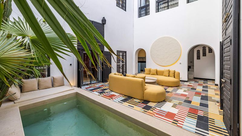 The courtyard seen through palm leaves: the pool, the curved mustard sofas, the multicoloured cement-tile floor and two arched doorways.