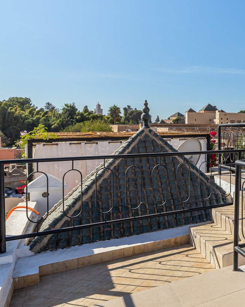 Vue depuis le rooftop : un toit pointu en tuiles vertes derrière le garde-corps en fer noir et, au loin, des palmiers et les toits de la médina.