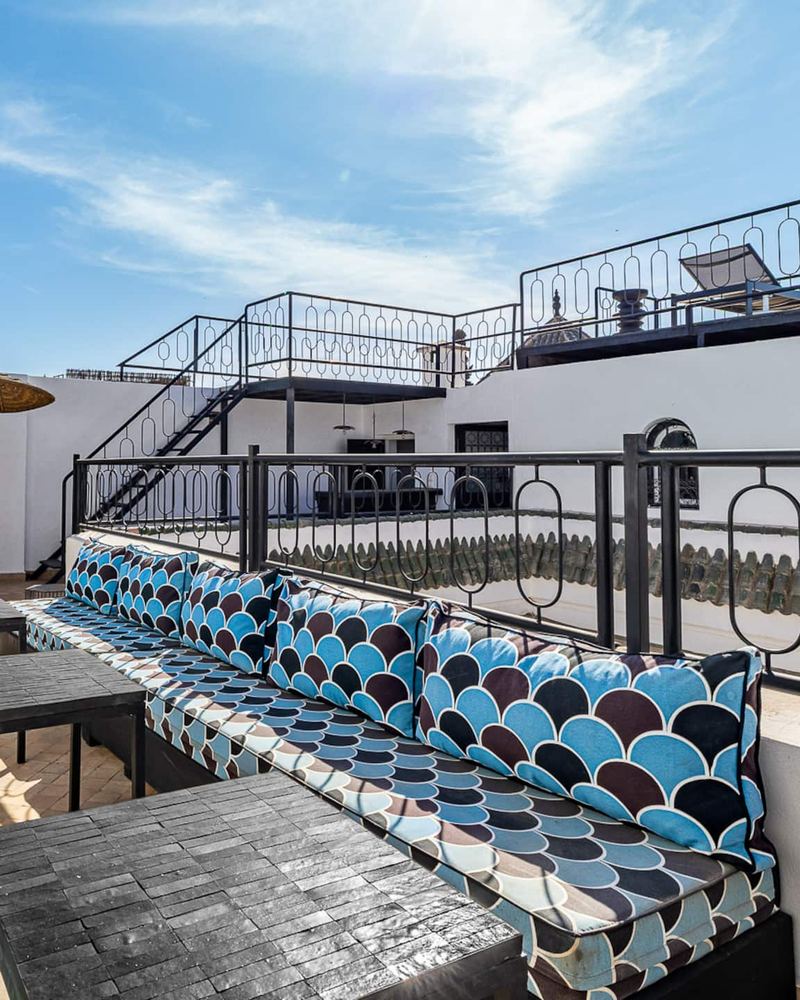 Une terrasse avec de longues banquettes aux coussins bleus et bruns, des tables basses noires et un escalier vers la terrasse du haut.
