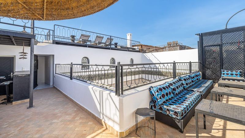 A roof terrace: sofas with blue and brown cushions, black low tables, iron railings and, higher up, sun loungers.