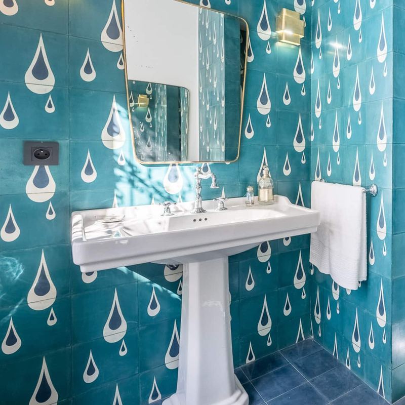 Une salle de bain aux carreaux bleu canard à motif de gouttes, avec un lavabo sur colonne et un miroir.