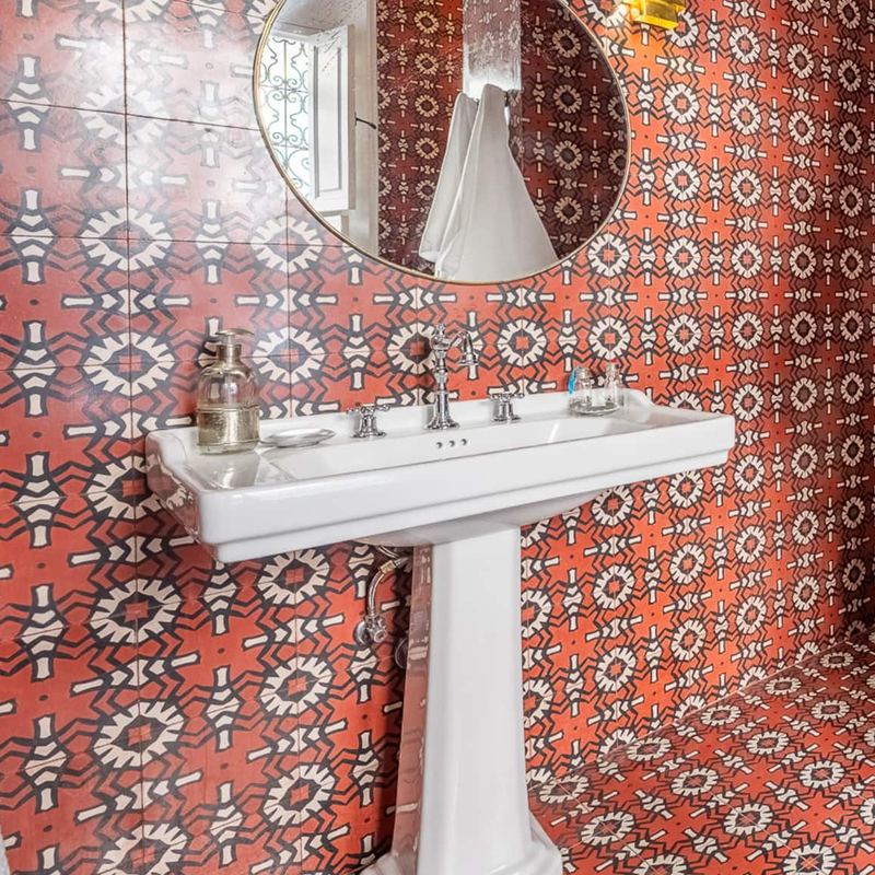 Une salle de bain aux carreaux rouges à motifs, avec un lavabo sur colonne et un miroir rond.